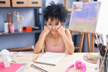 Young hispanic woman artist sitting on table with sad expression at art studio