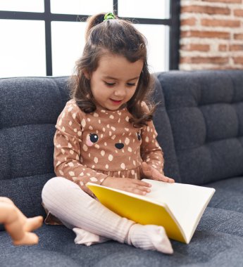 Adorable hispanic girl reading book sitting on sofa at home