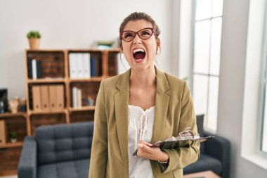 Young woman working at consultation office angry and mad screaming frustrated and furious, shouting with anger. rage and aggressive concept. 