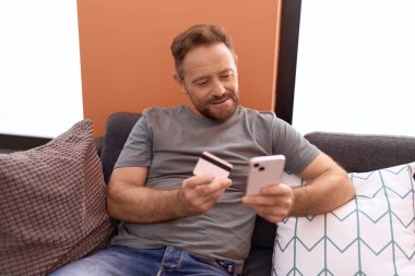 Evdeki koltukta oturan orta yaşlı bir adam akıllı telefon ve kredi kartı kullanıyor.