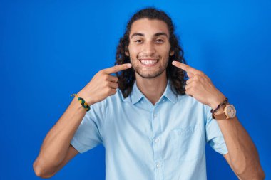 Mavi arka planda duran, neşeli gülümseyen ve parmaklarıyla ve ağzıyla işaret eden İspanyol genç adam. diş sağlığı kavramı. 