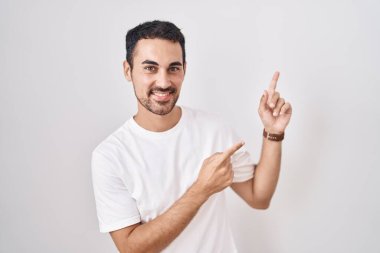 Handsome hispanic man standing over white background smiling and looking at the camera pointing with two hands and fingers to the side. 