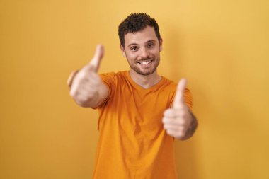 Young hispanic man standing over yellow background approving doing positive gesture with hand, thumbs up smiling and happy for success. winner gesture. 