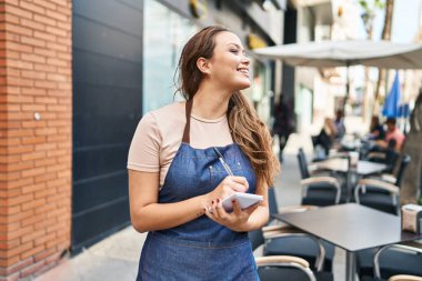 Young beautiful hispanic woman waitress smiling confident writing on notebook at coffee shop terrace