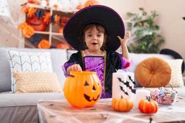 Adorable hispanic girl having halloween party putting sweets on pumpkin basket at home