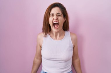 Brunette woman standing over pink background angry and mad screaming frustrated and furious, shouting with anger. rage and aggressive concept. 