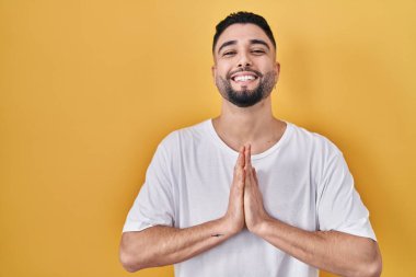 Young handsome man wearing casual t shirt over yellow background praying with hands together asking for forgiveness smiling confident. 