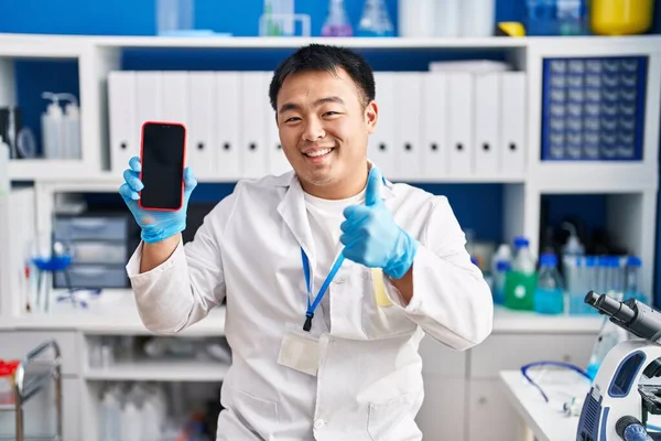Young chinese man working at scientist laboratory holding smartphone smiling happy and positive, thumb up doing excellent and approval sign