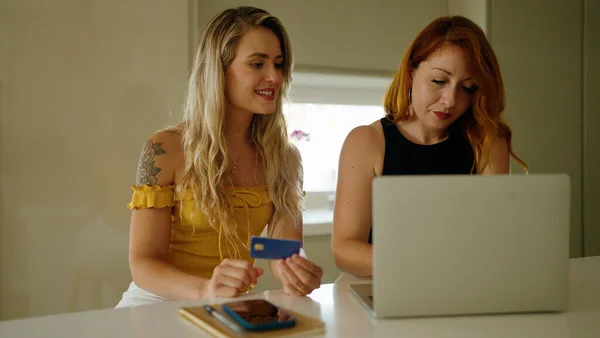 Two women using laptop and credit card sitting on table at kitchen