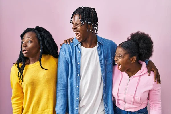 Group of three young black people standing together over pink background angry and mad screaming frustrated and furious, shouting with anger. rage and aggressive concept.