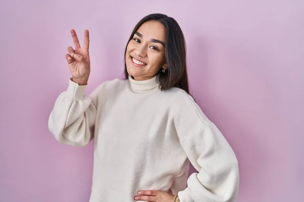 Young south asian woman standing over pink background smiling looking to the camera showing fingers doing victory sign. number two.