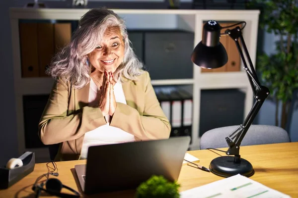 手で一緒に祈る夜遅くにコンピュータのラップトップを使用して作業灰色の髪を持つ中世の女性は自信を持って笑顔許しを求めて — ストック写真