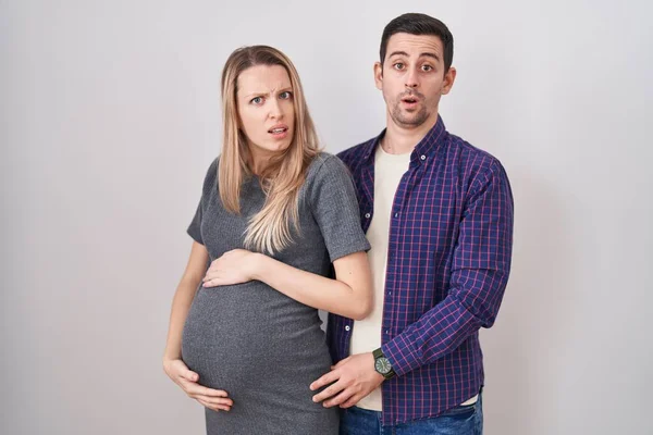 若いですカップル期待している赤ちゃんの上に立っている白い背景でショック顔 懐疑的かつ皮肉 驚きでオープン口 — ストック写真