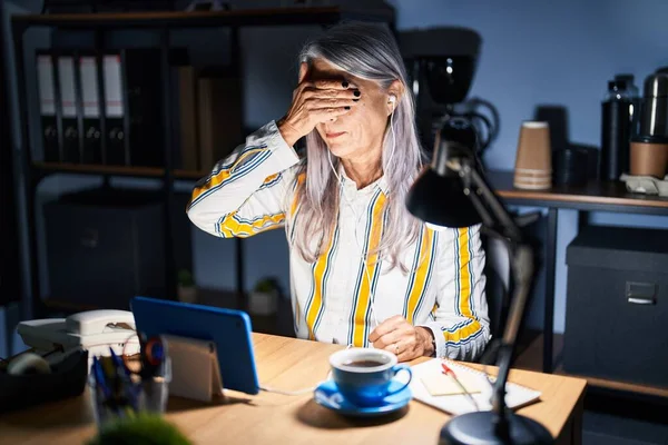 Middle age woman with grey hair working at the office at night smiling and laughing with hand on face covering eyes for surprise. blind concept.