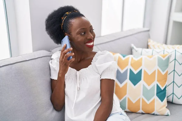 アフリカ系アメリカ人の女性が自宅でソファに座っているスマートフォンで話しています — ストック写真