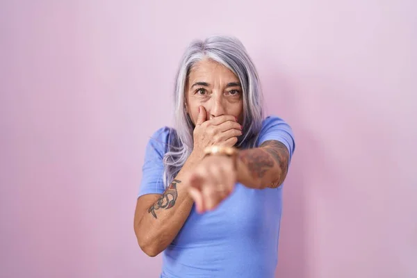 Middle age woman with tattoos standing over pink background laughing at you, pointing finger to the camera with hand over mouth, shame expression
