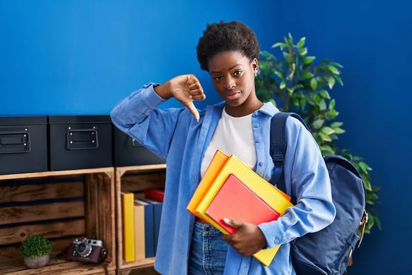 African american woman wearing student backpack and holding books with angry face, negative sign showing dislike with thumbs down, rejection concept