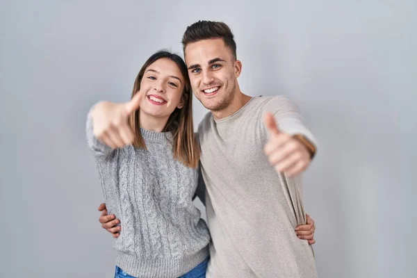 若いヒスパニック系のカップルは 手で積極的なジェスチャーを行うことを承認白い背景の上に立って 親指を笑顔と成功のために幸せ 勝者のジェスチャー — ストック写真