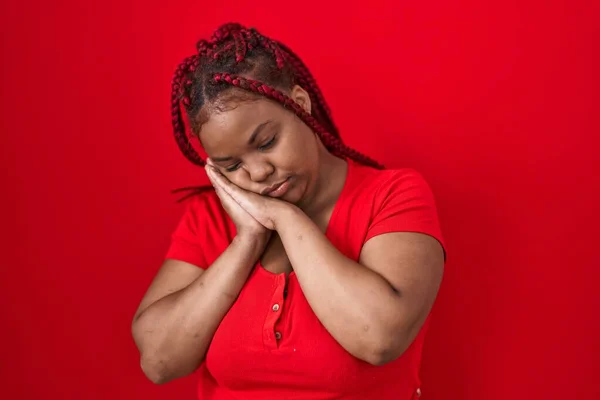 African american woman with braided hair standing over red background sleeping tired dreaming and posing with hands together while smiling with closed eyes.
