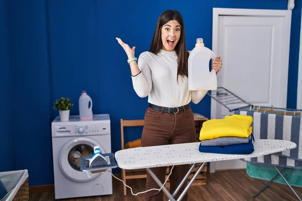 Young brunette woman with folded laundry after ironing celebrating victory with happy smile and winner expression with raised hands