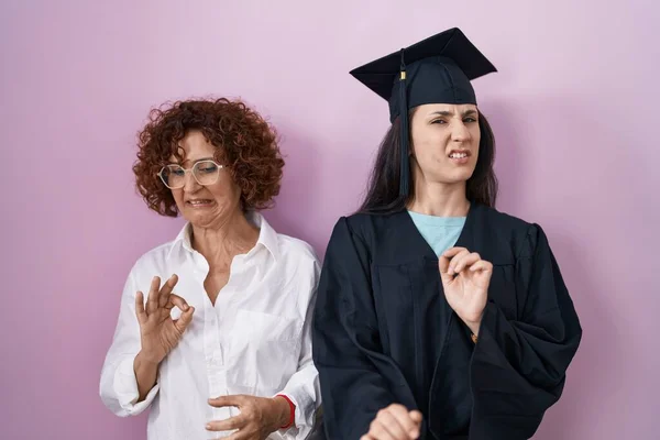 Hispanic mother and daughter wearing graduation cap and ceremony robe disgusted expression, displeased and fearful doing disgust face because aversion reaction. with hands raised