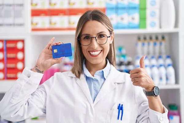 Young woman working at pharmacy drugstore holding credit card smiling happy and positive, thumb up doing excellent and approval sign