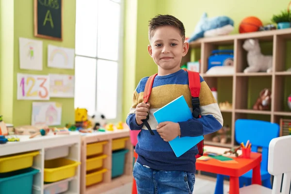 Adorable hispanic boy student wearing backpack holding book at kindergarten