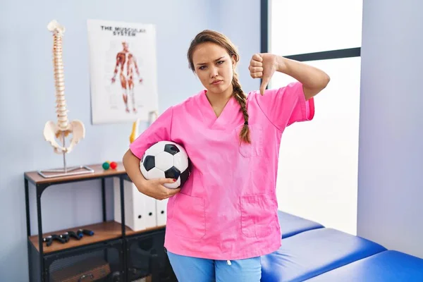 Young Hispanic Woman Working Physiotherapy Clinic Holding Football Ball Angry — Foto de Stock