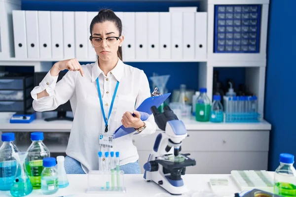 Young brunette woman working at scientist laboratory pointing down looking sad and upset, indicating direction with fingers, unhappy and depressed.