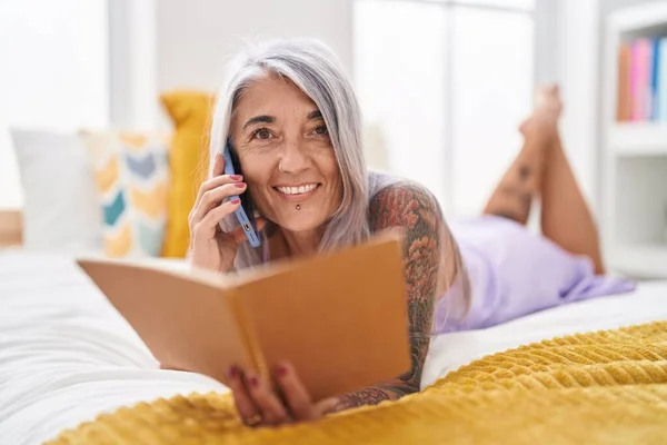 Middle age grey-haired woman talking on smartphone reading book at bedroom