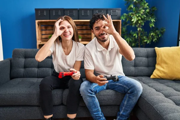 Young couple playing video game holding controller at home smiling happy doing ok sign with hand on eye looking through fingers