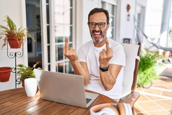 Middle age man using computer laptop at home showing middle finger doing fuck you bad expression, provocation and rude attitude. screaming excited