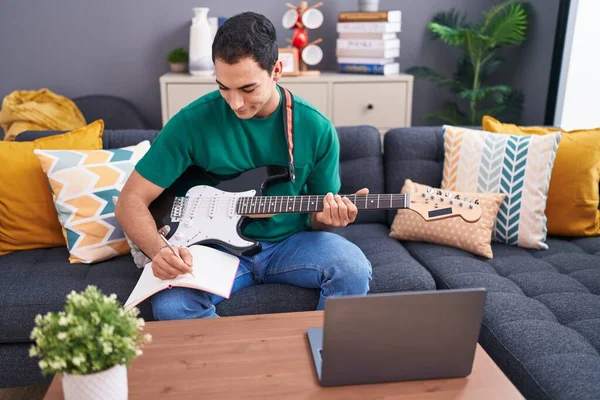 Young hispanic man having online electrical guitar class sitting on sofa at home