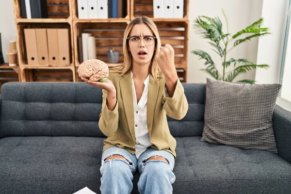 Young blonde woman working at therapy office holding brain annoyed and frustrated shouting with anger, yelling crazy with anger and hand raised