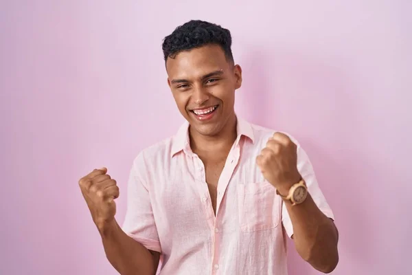 ピンクの背景に立っている若いヒスパニック系の男性は非常に満足し 腕を上げて勝者のジェスチャーを行う興奮し 笑顔と成功のために叫んでいます お祝いのコンセプト — ストック写真
