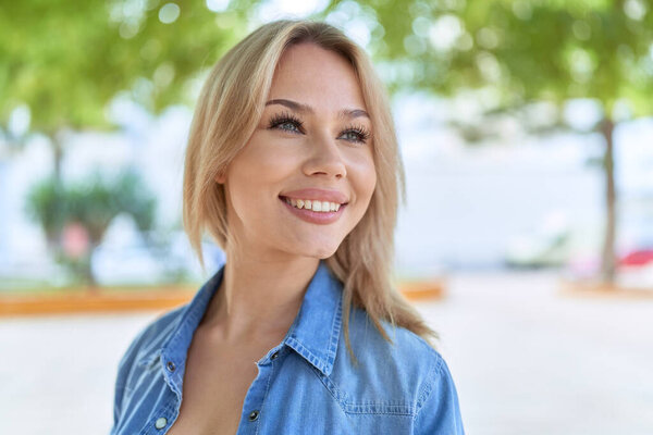 Молодая блондинка с уверенной улыбкой смотрит в сторону парка