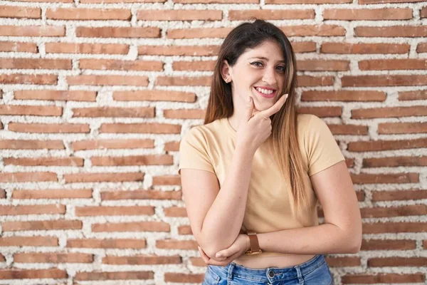 Young brunette woman standing over bricks wall looking confident at the camera smiling with crossed arms and hand raised on chin. thinking positive.