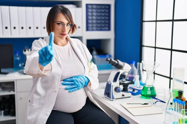 Pregnant woman working at scientist laboratory pointing with finger up and angry expression, showing no gesture