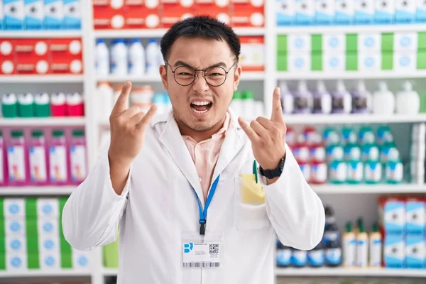 Chinese young man working at pharmacy drugstore shouting with crazy expression doing rock symbol with hands up. music star. heavy music concept.