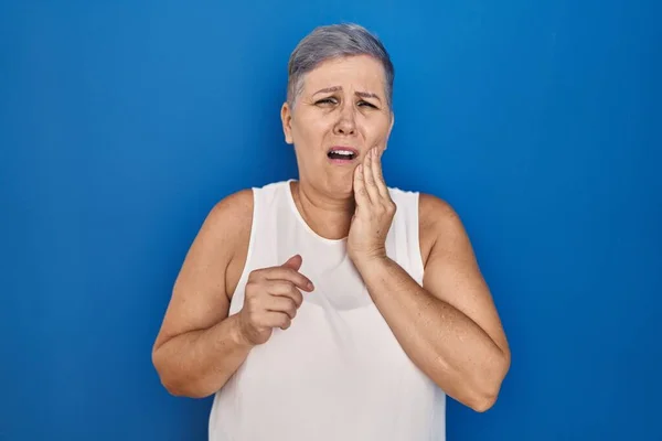 中年白人妇女站在蓝色的背景上 手部有疼痛的表情 手部因牙痛或牙齿疾病而接触嘴巴 — 图库照片