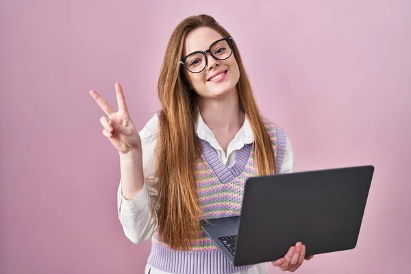年轻的高加索女人在使用电脑笔记本电脑工作时 满脸喜色地微笑着 用手指对着相机做胜利的标志眨眼 第二点 — 图库照片