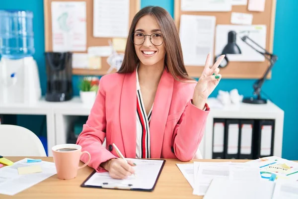 オフィスで働く若いヒスパニック系の女性は カメラでの勝利サインを指で行うと 幸せな顔のウィンクで笑顔眼鏡をかけています — ストック写真