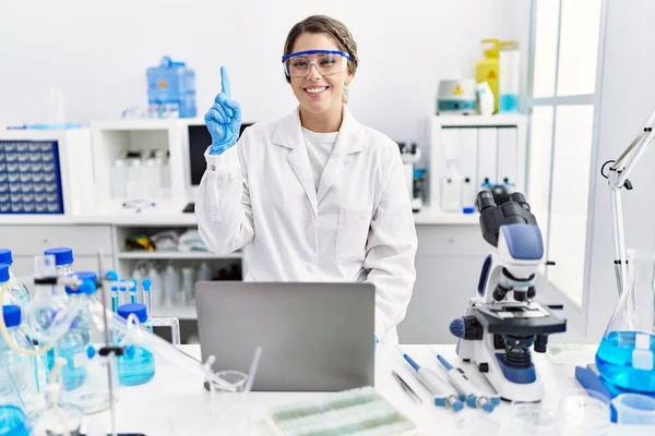 Young hispanic woman wearing scientist uniform working at laboratory surprised with an idea or question pointing finger with happy face, number one