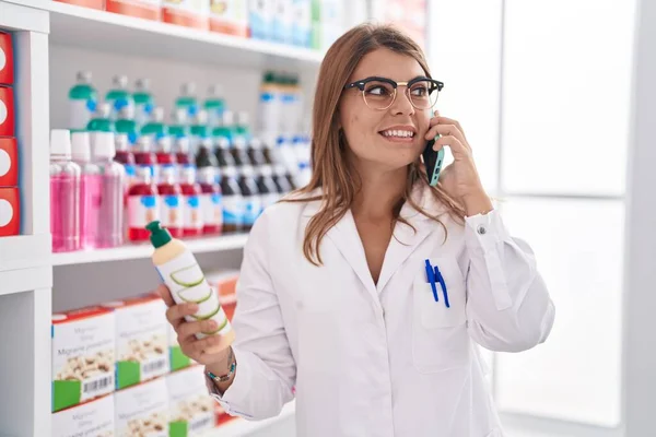 Молодая Женщина Фармацевт Держит Бутылку Шампуня Разговаривая Смартфоне Аптеке — стоковое фото