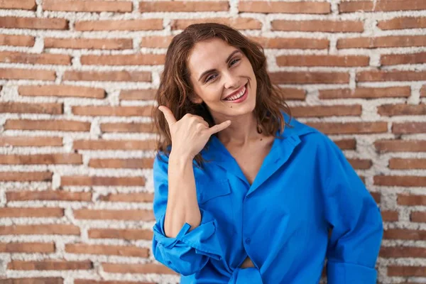 Beautiful brunette woman standing over bricks wall smiling doing phone gesture with hand and fingers like talking on the telephone. communicating concepts.