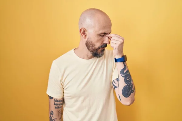 Hispanic man with tattoos standing over yellow background tired rubbing nose and eyes feeling fatigue and headache. stress and frustration concept.
