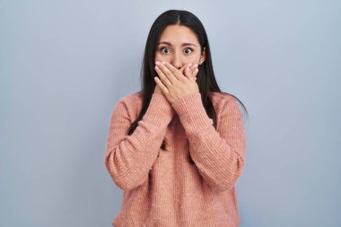 Young latin woman standing over blue background shocked covering mouth with hands for mistake. secret concept. 
