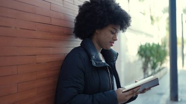 African american woman reading book leaning on wall at street