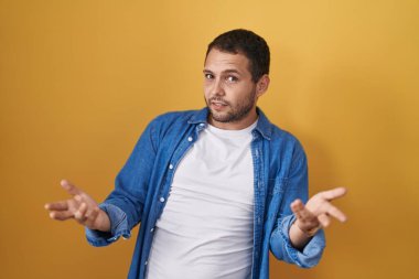 Hispanic man standing over yellow background clueless and confused with open arms, no idea concept. 