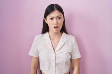 Chinese young woman standing over pink background in shock face, looking skeptical and sarcastic, surprised with open mouth 